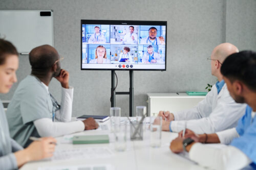 Group of doctors sitting at table and looking at big screen display of an online meeting with colleagues