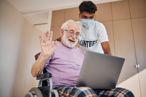 Happy elderly man in wheelchair participates in a video call on a laptop with a volunteer escort by his side