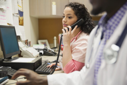 Female medical professional using telephone while working at desktop computer with colleague in foreground