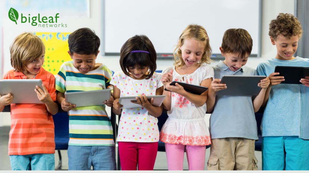 A group of six cheerful children, with diverse ethnic backgrounds, are each holding tablets and expressing excitement in a classroom setting.