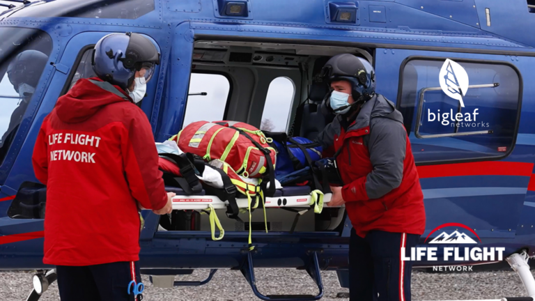 Emergency medical service personnel in red uniforms are transferring a patient on a stretcher into a blue Life Flight Network helicopter.