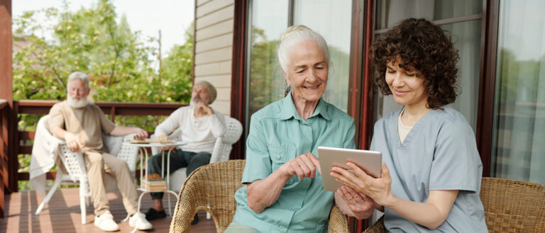 Elderly woman with caregiver learning to use a tablet on a sunny assisted living facility porch, with senior men conversing in the background.
