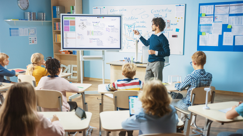 Teacher using an interactive digital whiteboard to explain a lesson in a modern classroom filled with diverse students attentively watching.
