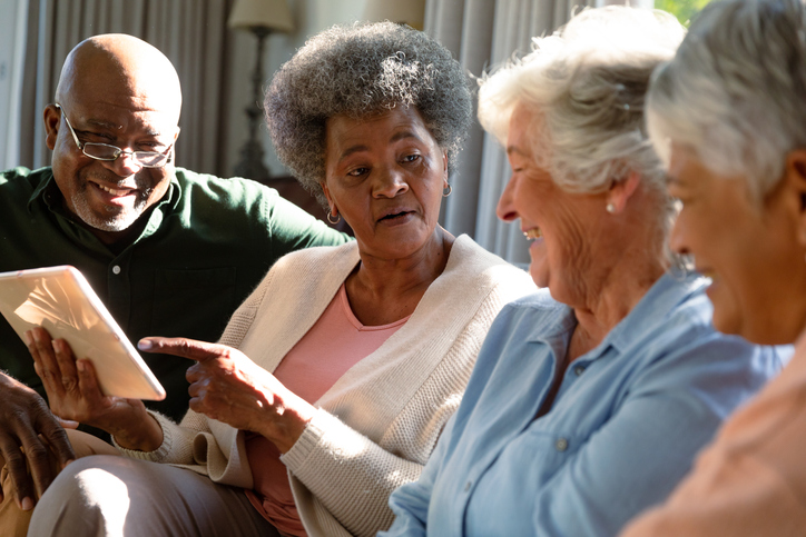 Photo of three happy diverse senior women and african american male friend sitting on sofa using a tablet