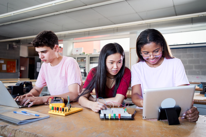 Three high school students engaged in a STEM project, using laptops and electronic circuit boards in a lab setting.