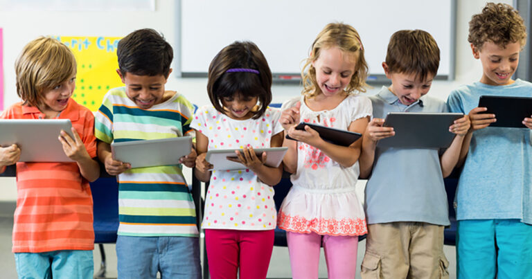 Group of 6 young elementary aged students standing at the front of a classroom, each holding an iPad and looking down at it.